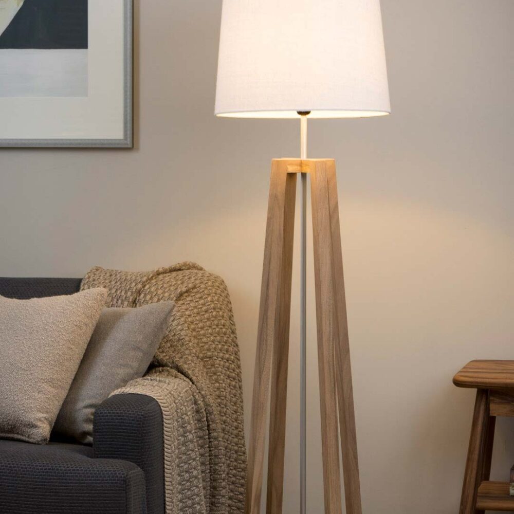 A modern tripod floor lamp with a white drum shade and natural wood legs, shown lit in a cozy living room setting next to a sofa.