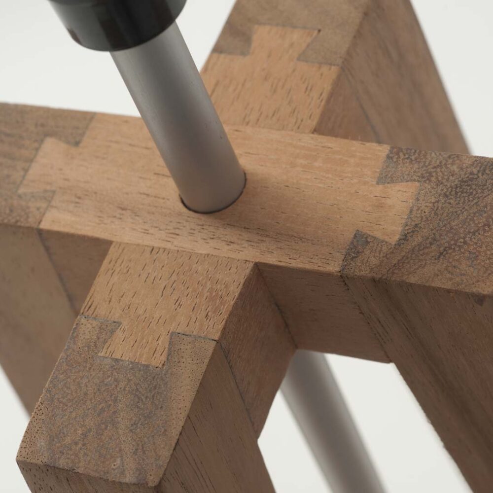 Close-up of a wooden lamp with interlocking (tenon-joint) style wood pieces and a silver metal rod passing through the center, shown against a plain light background.