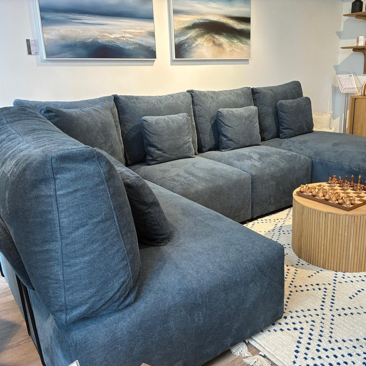 A large L-shaped sectional sofa upholstered in textured slate blue fabric, styled in a modern living room setting with a round wooden coffee table, patterned rug, and wall art.