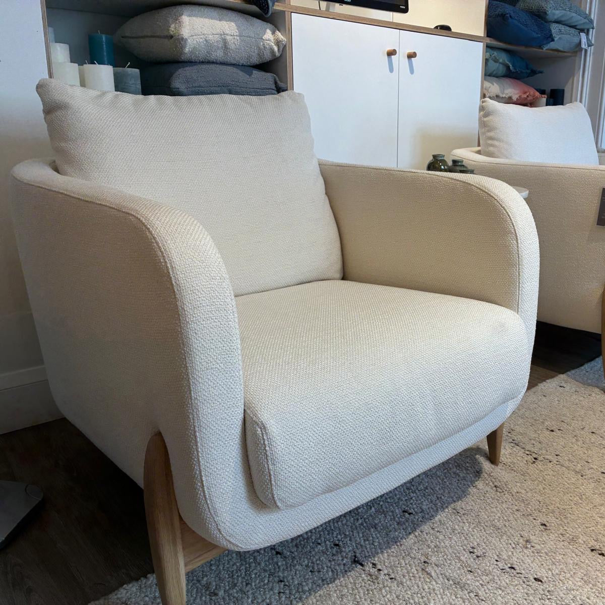 A cream-colored upholstered armchair with light wood legs, shown in a home interior setting next to another matching armchair.