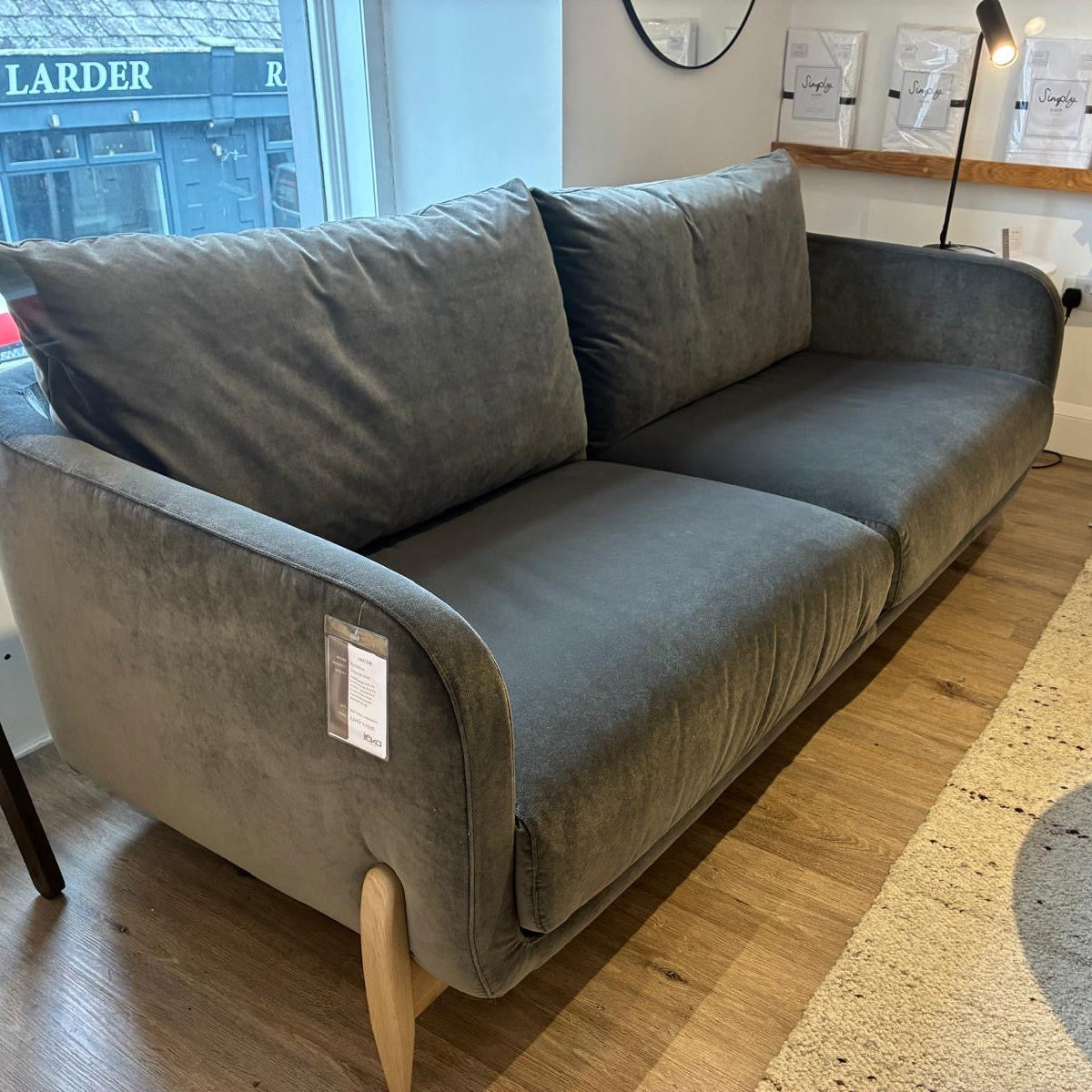A gray velvet loveseat sofa with light wood legs, shown in a home showroom setting with a marble coffee table and wooden floor.