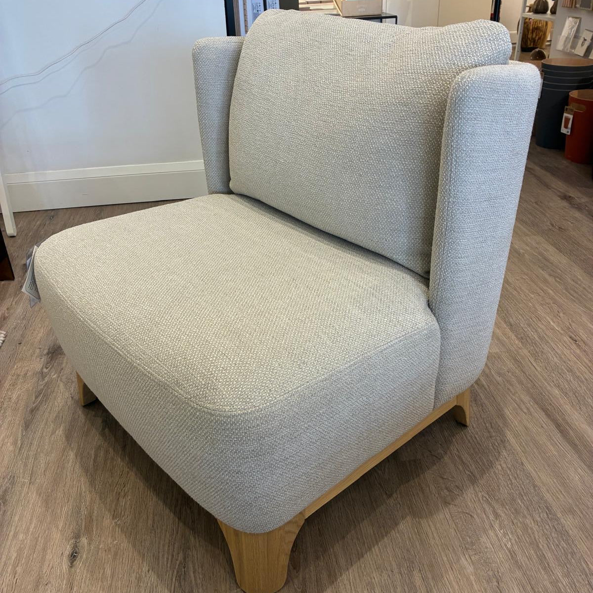 A light beige textured fabric armchair with light wood legs, photographed in a furniture showroom on a wood-look floor.