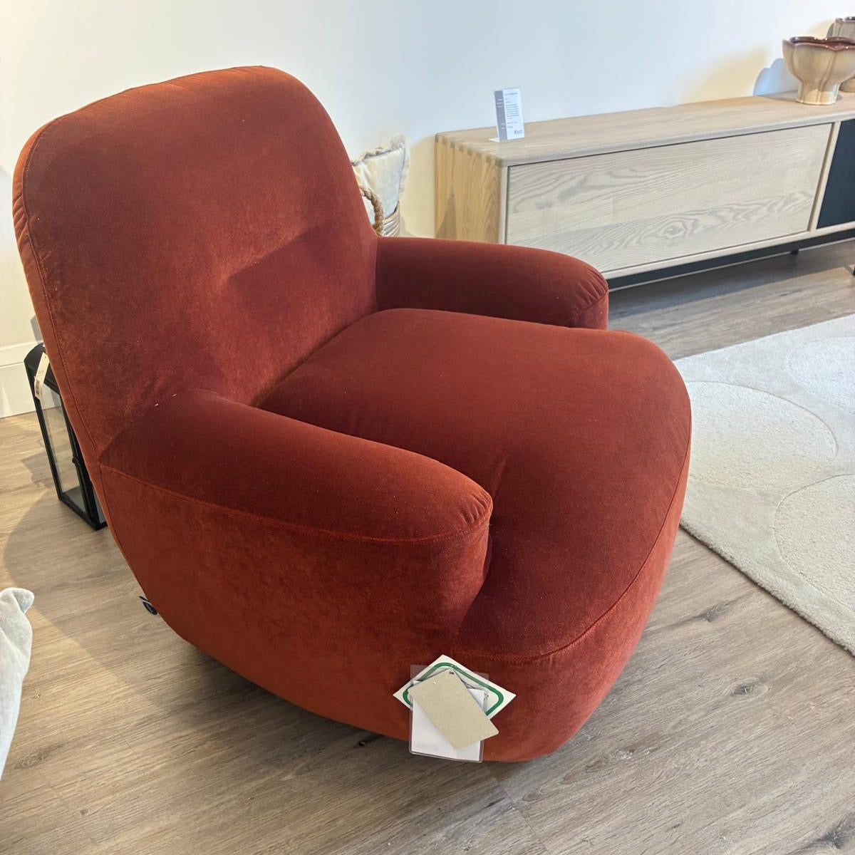 A plush, rounded accent chair upholstered in a textured rust-orange velvet fabric, shown in a modern living room setting with a light wood floor and a light-colored cabinet in the background.
